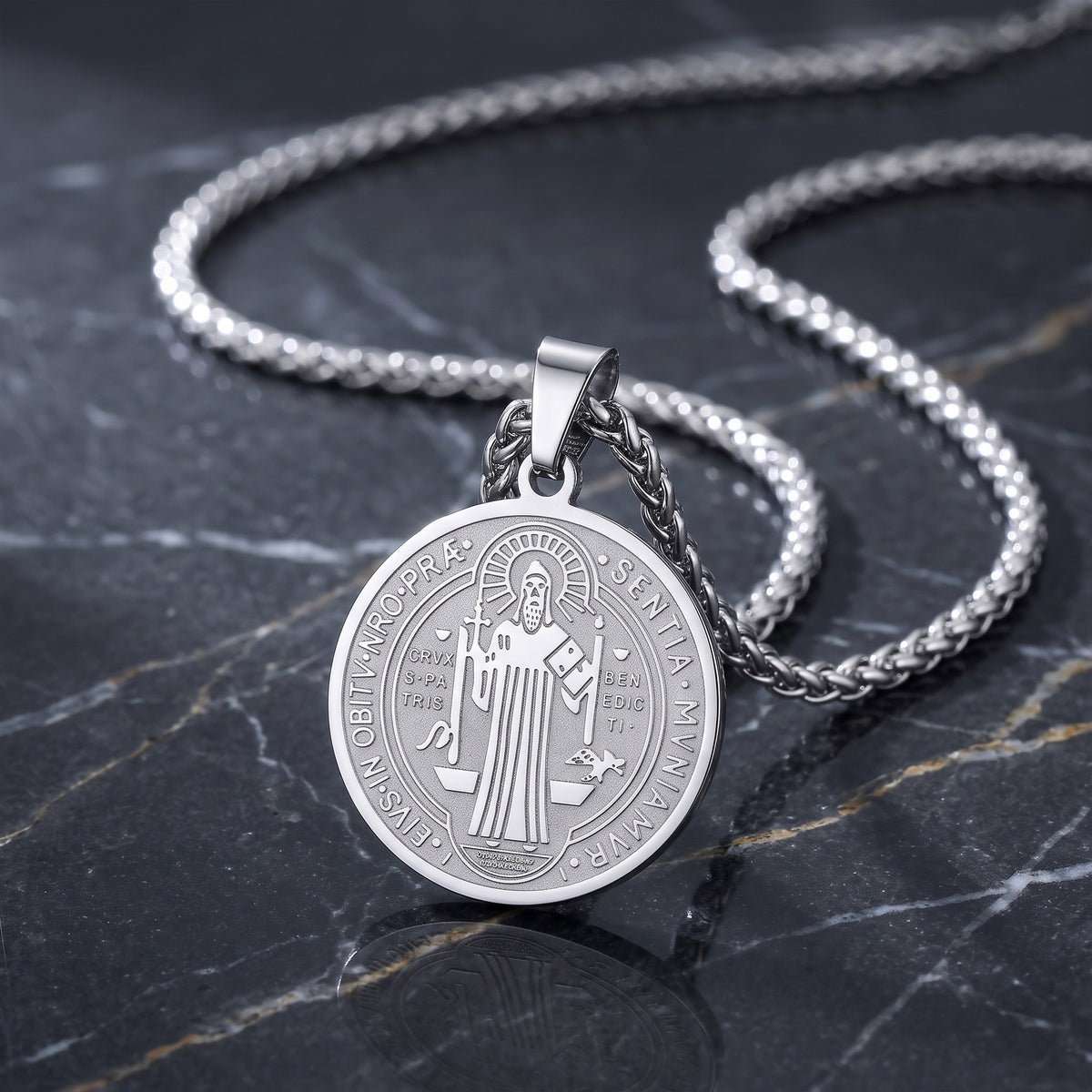 A silver round pendant necklace featuring an engraved religious figure, displayed on a dark marble surface with a matching silver chain.