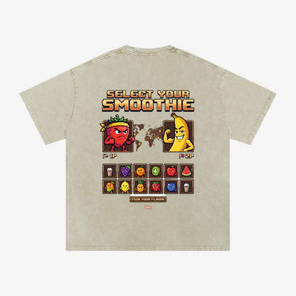 A light beige graphic t-shirt shown from the back, featuring a retro video game style 'Select Your Smoothie' design with pixel art characters and a grid of colorful smoothie icons.