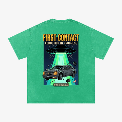 A green short-sleeve t-shirt with a graphic print on the back. The graphic features a gray Cadillac Escalade SUV under a glowing green UFO with the text 'FIRST CONTACT ABDUCTION IN PROGRESS' in yellow and white.