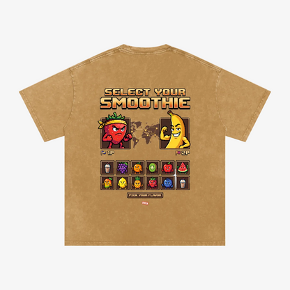 A tan short-sleeve t-shirt with a retro video game style graphic on the back. The graphic features the text 'SELECT YOUR SMOOTHIE' above pixel-art characters representing fruit smoothie flavors (Strawberry, Pineapple, Mango, etc.