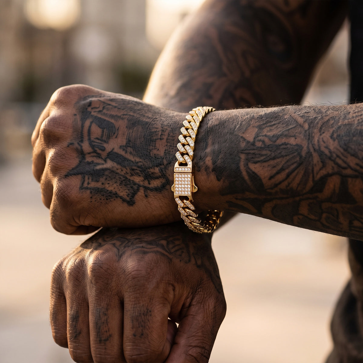 A close-up lifestyle shot of a man's tattooed arms crossed, showcasing a thick gold chain bracelet with a rectangular diamond-encrusted bezel clasp.