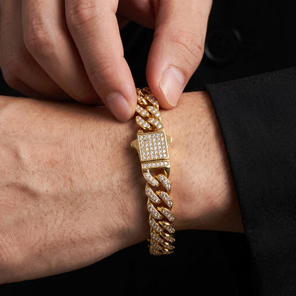 A close-up of a gold-toned Cuban link bracelet with pavé-set clear stones, worn on a person's wrist while another hand adjusts it. The background is dark, and the person is wearing a black suit jacket.