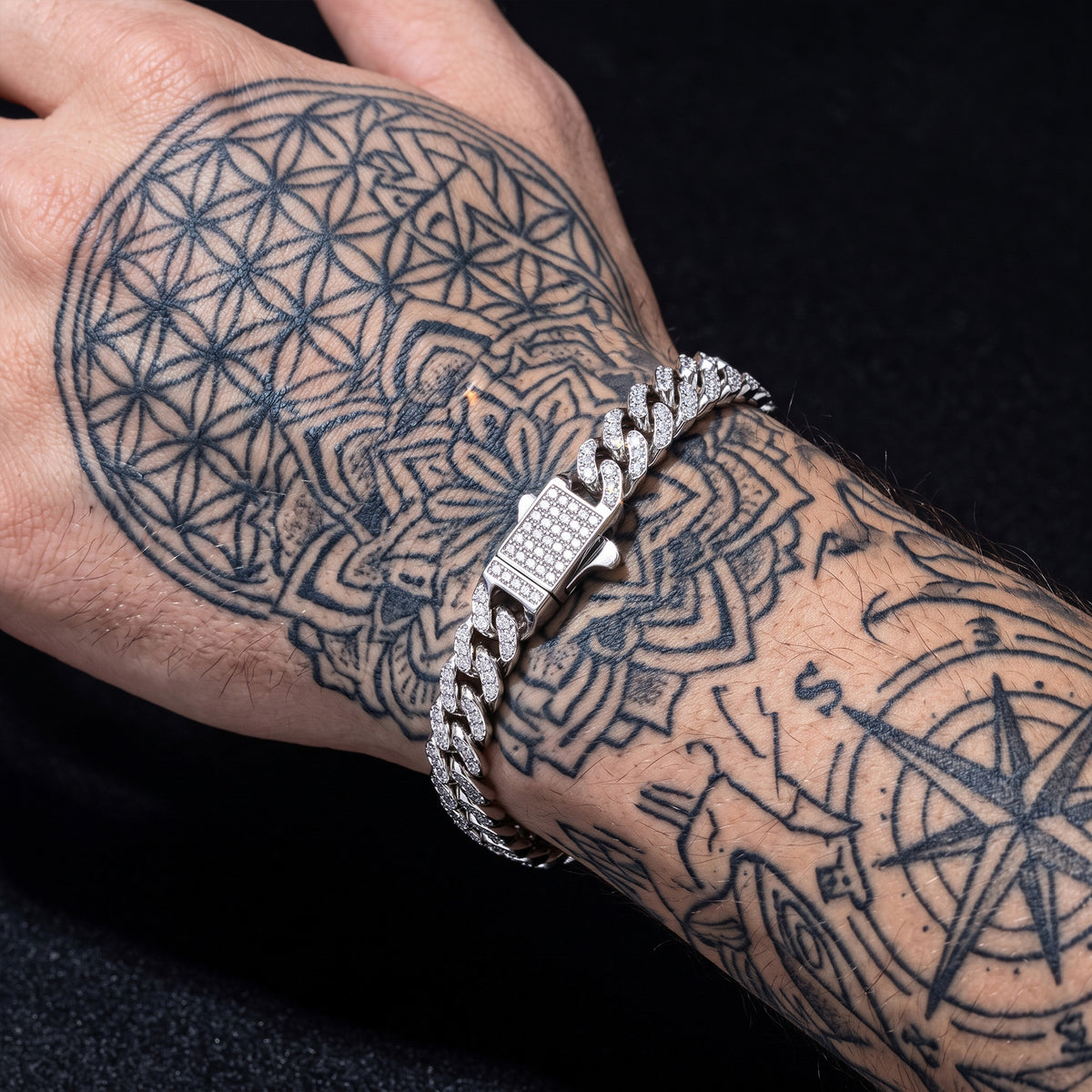A close-up of a tattooed wrist wearing a silver-toned Cuban link bracelet paved with small clear stones, set against a black background.
