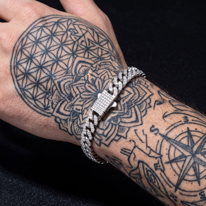 A close-up of a tattooed wrist wearing a silver-toned Cuban link bracelet paved with small clear stones, set against a black background.