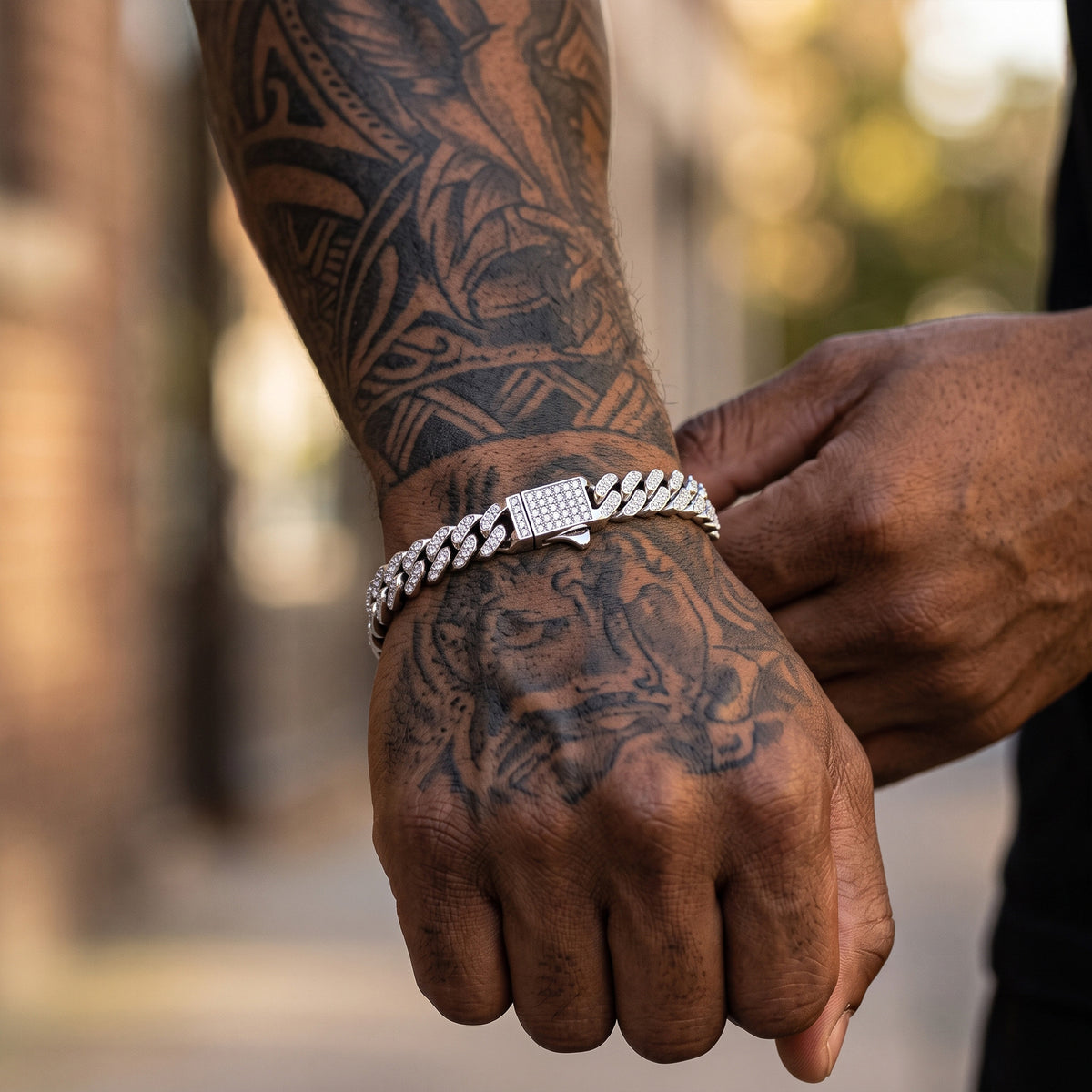 A close-up lifestyle shot of a man's tattooed wrist wearing a silver Cuban link chain bracelet with a diamond-encrusted clasp, with another hand adjusting the chain.
