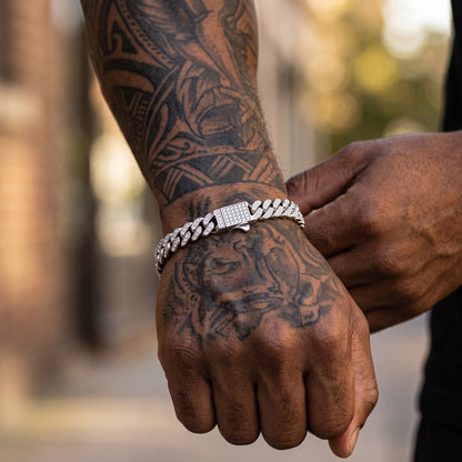 A close-up lifestyle shot of a man's tattooed wrist wearing a silver Cuban link chain bracelet with a diamond-encrusted clasp, with another hand adjusting the chain.