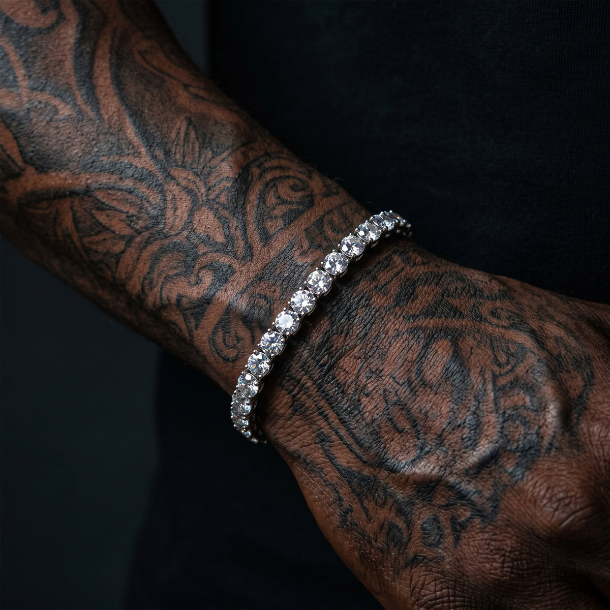 A close-up of a silver-toned tennis bracelet with clear stones worn on a tattooed wrist against a dark background.
