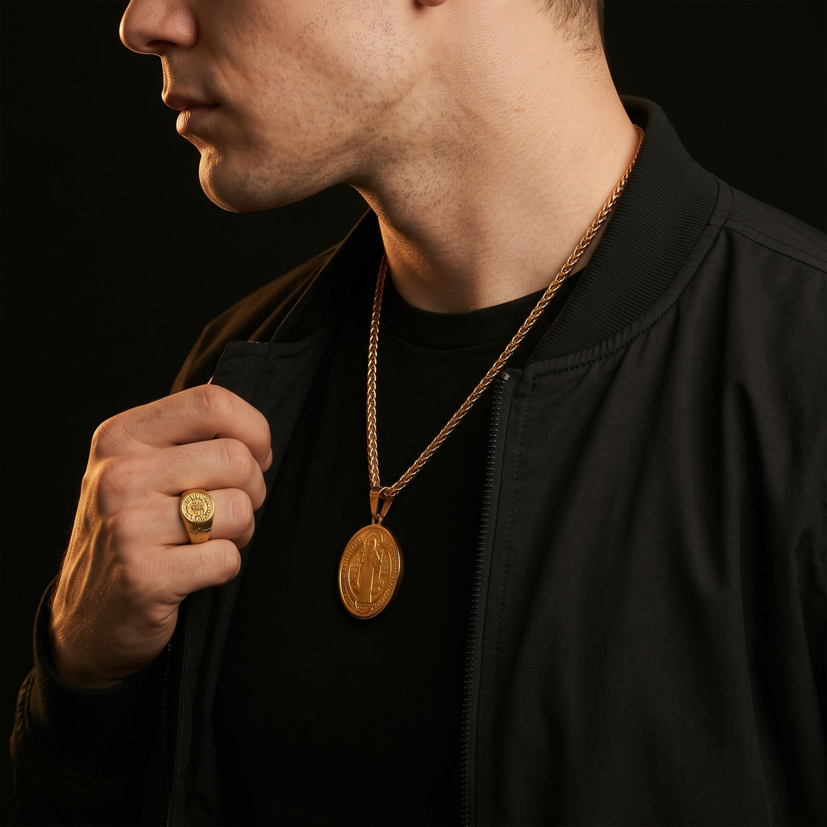 A close-up of a man wearing a gold pendant necklace with an oval religious medallion, styled with a gold ring on his finger and a black bomber jacket against a dark background.