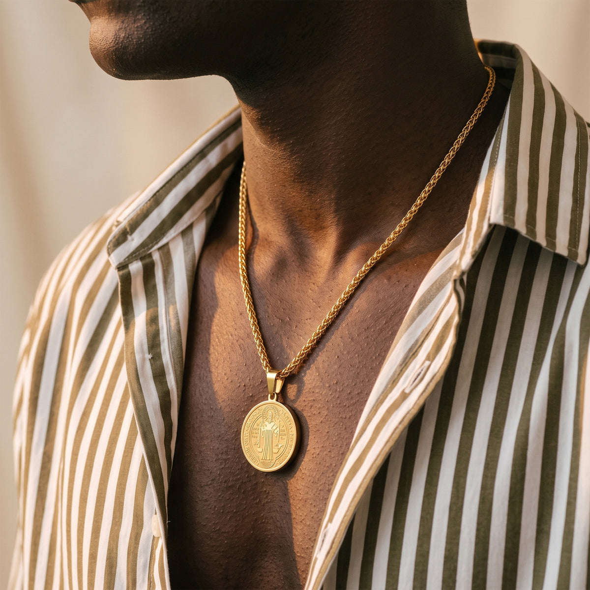 A close-up lifestyle shot of a man wearing a gold pendant necklace with a circular pendant featuring a cross, styled with a striped button-down shirt.