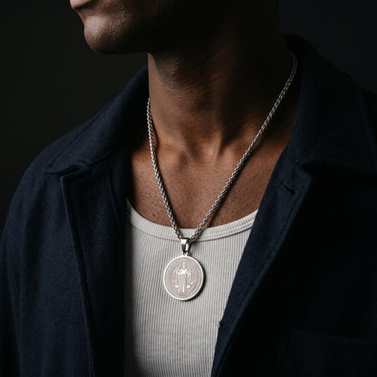 A close-up of a man wearing a silver pendant necklace with a cactus design, styled with a dark navy overshirt and a light gray ribbed t-shirt against a dark background.