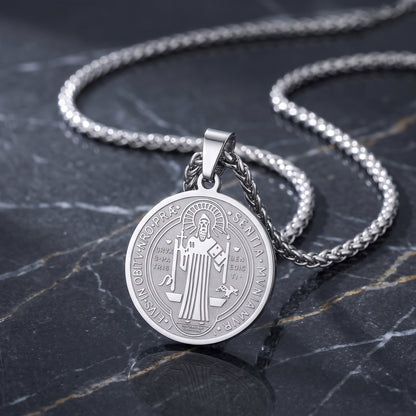 A silver round pendant necklace featuring an engraved religious figure, displayed on a dark marble surface with a matching silver chain.