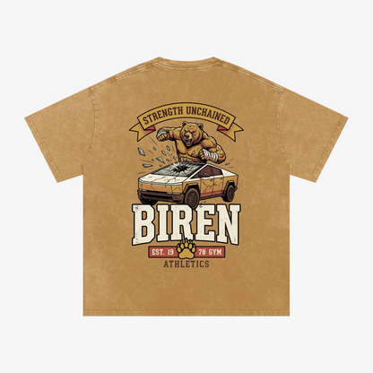 A tan short-sleeve t-shirt with a large graphic print on the back. The graphic features a cartoon bear crushing a car with the text 'STRENGTH UNCHAINED' above and 'BIREN' below, along with 'EST.