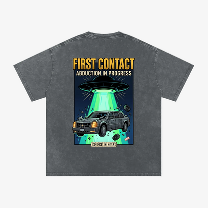 A dark gray acid-wash t-shirt with a large graphic print on the back. The graphic features a gray Cadillac SUV under a glowing green UFO beam with the text 'FIRST CONTACT ABDUCTION IN PROGRESS' in yellow and white.