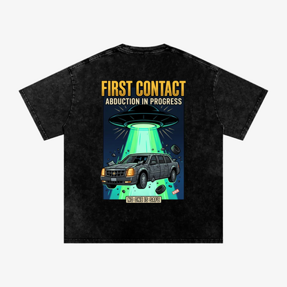 A black acid-wash graphic t-shirt shown from the back, featuring a cartoon illustration of a gray SUV being abducted by a UFO with a green beam of light, and bold yellow and white text reading 'FIRST CONTACT ABDUCTION IN PROGRESS' and 'NOT ALL IS GAIN'.