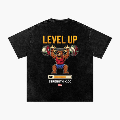 A black acid-wash graphic t-shirt featuring a cartoon bear lifting a barbell with the text 'LEVEL UP' and 'STRENGTH +100' on the back, displayed against a light gray background.