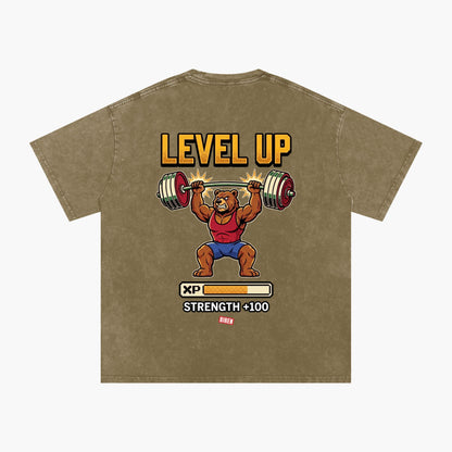 A brown mineral-washed t-shirt featuring a retro-style graphic of a bear lifting a barbell with the text 'LEVEL UP' and 'STRENGTH +100 XP'.