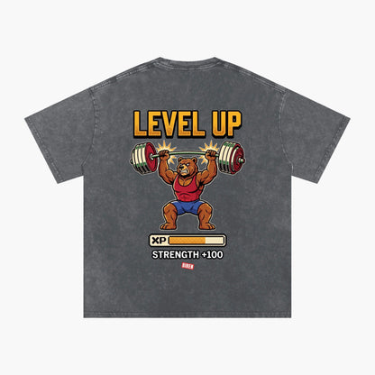 A dark gray acid-wash t-shirt featuring a cartoon graphic of a bear lifting a barbell under the text 'LEVEL UP' and above an XP progress bar graphic.