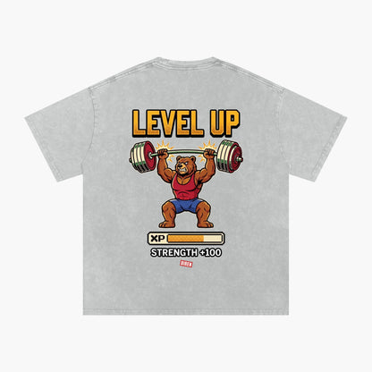 A light gray heathered t-shirt featuring a retro video game 'Level Up' graphic of a cartoon bear lifting a barbell, with 'LEVEL UP' in yellow text and a progress bar graphic below.