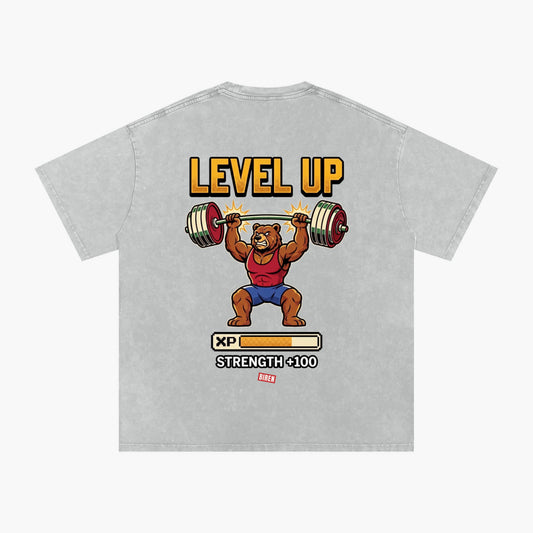 A light gray heathered t-shirt featuring a retro video game 'Level Up' graphic of a cartoon bear lifting a barbell, with 'LEVEL UP' in yellow text and a progress bar graphic below.